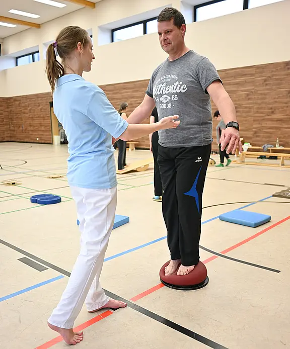 Physiotherapeutin unterstützt einen Mann beim Gleichgewichtstraining: Er steht barfuß auf einem wackeligen Balancekissen und hebt das andere Bein, Arme seitlich ausgestreckt. Die Übung findet in einer Sporthalle statt.