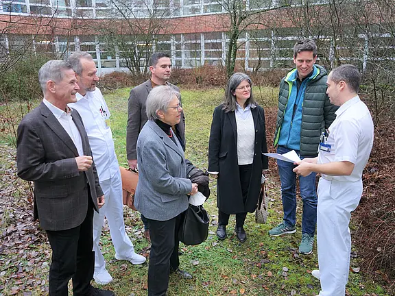 Sieben Personen stehen im Freien vor einem Gebäude. Zwei tragen weiße Klinikbekleidung. Eine Person in Weiß zeigt ein Blatt Papier und erklärt etwas. Die anderen hören zu. Die Szene wirkt wie ein kurzes Gespräch oder eine Besprechung in einem Krankenhausumfeld.