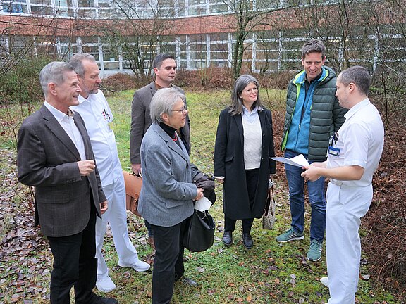 Sieben Personen stehen im Freien vor einem Gebäude. Zwei tragen weiße Klinikbekleidung. Eine Person in Weiß zeigt ein Blatt Papier und erklärt etwas. Die anderen hören zu. Die Szene wirkt wie ein kurzes Gespräch oder eine Besprechung in einem Krankenhausumfeld.