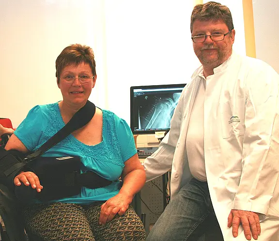 Patientin mit Schulterorthese und Armschlinge sitzt neben einem Arzt im weißen Kittel. Beide lächeln. Hinter ihnen zeigt ein Monitor ein Röntgenbild der Schulter. Szene wirkt wie eine Nachuntersuchung in der Praxis.