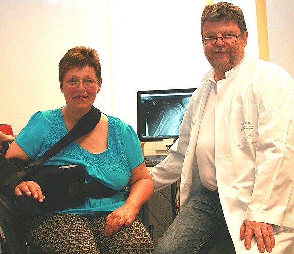 Patientin mit Schulterorthese und Armschlinge sitzt neben einem Arzt im weißen Kittel. Beide lächeln. Hinter ihnen zeigt ein Monitor ein Röntgenbild der Schulter. Szene wirkt wie eine Nachuntersuchung in der Praxis.