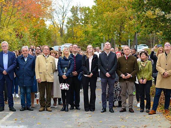 Eine größere Gruppe Erwachsener steht dicht beieinander im Freien vor herbstlichen Bäumen. Sie blicken konzentriert nach oben, als würden sie einer Vorführung oder Ansprache folgen. Vorn stehen mehrere Personen in Jacken und Mänteln; hinter ihnen weitere Zuschauer.