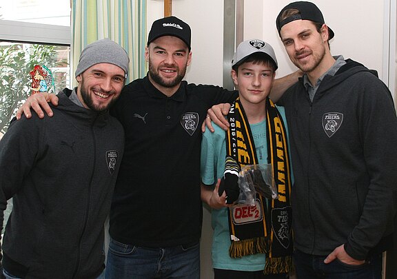 Drei erwachsene Männer in grauer Teamkleidung posieren lächelnd mit einem Jungen, der Fan-Schal und Kappe trägt. Einer legt den Arm um ihn. Die Szene wirkt wie ein freundliches Treffen zwischen Sportlern und Fan.