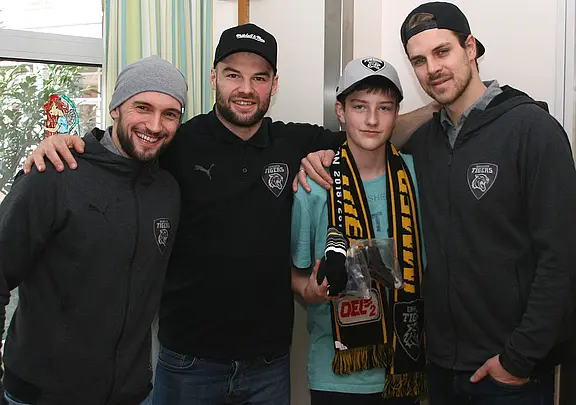 Drei erwachsene Männer in grauer Teamkleidung posieren lächelnd mit einem Jungen, der Fan-Schal und Kappe trägt. Einer legt den Arm um ihn. Die Szene wirkt wie ein freundliches Treffen zwischen Sportlern und Fan.