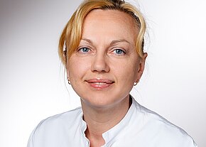 Tanja Dick Ein Lächeln einer Frau mit blonden Haaren, die in einem weißen Laborkittel gekleidet ist. Sie hat blaue Augen und trägt kleine Ohrringe. Der Hintergrund ist hell und neutral gehalten.