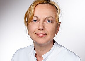 Tanja Dick Ein Lächeln einer Frau mit blonden Haaren, die in einem weißen Laborkittel gekleidet ist. Sie hat blaue Augen und trägt kleine Ohrringe. Der Hintergrund ist hell und neutral gehalten.