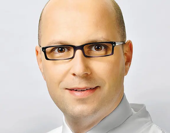 Porträt eines lächelnden Mannes mit Glatze und dunkler Brille vor neutralem Hintergrund. Nahaufnahme des Gesichts, direkter Blick in die Kamera, seriöser Ausdruck, Business-Hemd.