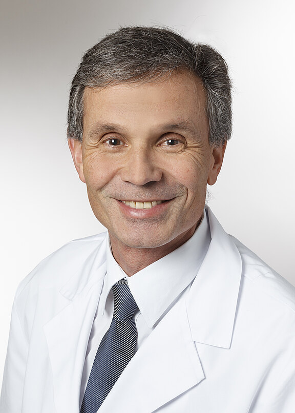 Porträt eines lächelnden, grauhaarigen Mannes in weißem Arztkittel und gestreifter Krawatte vor hellem Hintergrund, der direkt in die Kamera blickt.