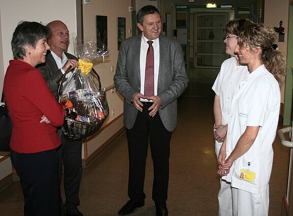 Im Krankenhausflur stehen drei Besucher zwei Pflegekräften in weißen Kitteln gegenüber. Alle lächeln und unterhalten sich. Einer der Besucher hält einen großen Geschenkkorb, offenbar als Dank oder Gruß für das Team.