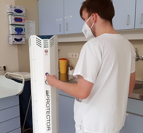 Ein medizinisches Fachpersonal steht in einem Behandlungsraum und bedient ein Gerät mit der Aufschrift "AIR PROTECTOR". Das Gerät hat eine schlanke, weiße Form und ist auf einer Fläche platziert, während im Hintergrund Küchenschränke und Utensilien sichtbar sind.