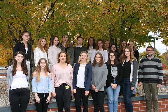 Gruppenfoto von rund 20 jungen Erwachsenen in zwei Reihen, entspannt lächelnd. Sie stehen draußen vor dichtem, gelbgrünem Herbstlaub. Freizeit- bis Business-Casual-Kleidung; rechts steht ein Mann mit Brille und Bart. Der Fokus liegt auf Gemeinschaft und Teamgefühl.