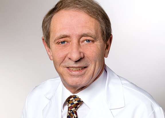 Porträt eines älteren Mannes in weißem Kittel und gemusterter Krawatte, leicht lächelnd, schaut direkt in die Kamera vor neutralem Hintergrund.