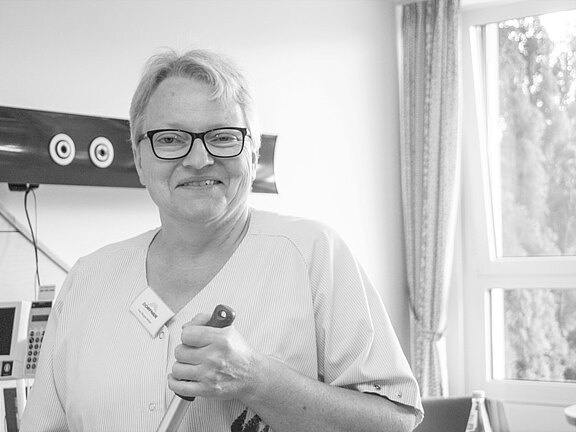 Schwarzweißfoto: lächelnde Person mit Brille und Namensschild in Arbeitskleidung, in einem Klinik- oder Pflegezimmer, hält einen Handgriff. Im Hintergrund stehen Geräte und ein Fenster.