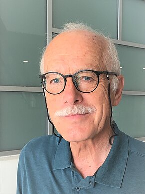 Klaus Pietruska Porträt eines älteren Mannes mit weißem Schnurrbart und runder Brille mit Band. Er trägt ein blaugrünes Polohemd, blickt zur Seite. Neutraler Innenhintergrund.
