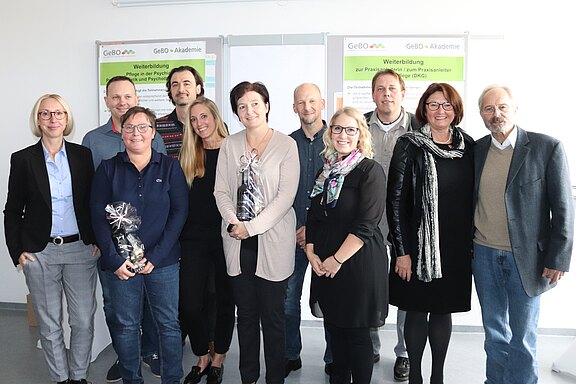 Eine Gruppe von zehn Personen steht zusammen in einem Raum. Sie lächeln und halten Geschenke in den Händen. Im Hintergrund sind Plakate zur Weiterbildung in der Pflege zu sehen. Die Atmosphäre wirkt freundlich und feierlich.