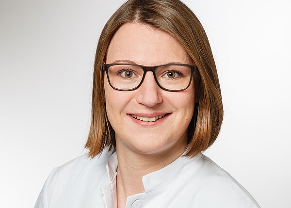 Porträt einer lächelnden Person mit schulterlangen braunen Haaren und dunkler Brille, in weißem Kittel, vor neutral hellem Hintergrund, direkt in die Kamera blickend.