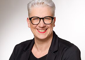 Anke Friedrich Porträtfoto einer lächelnden Person mit kurzem weißem Haar, schwarzer Brille und rotem Lippenstift, in schwarzer Jacke vor hellem, neutralem Hintergrund; Blick frontal in die Kamera.