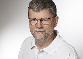 Prof. Dr. med. Rainer Abel Porträt eines Mannes mit grauen Haaren und Brille. Er trägt ein weißes Polo-Shirt und hat einen freundlichen Gesichtsausdruck. Der Hintergrund ist hell und neutral.
