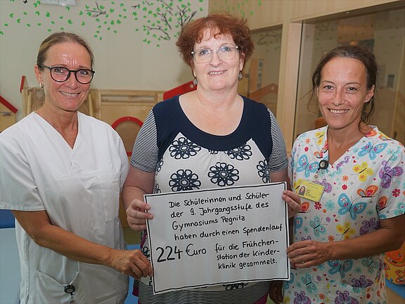 Drei Frauen, zwei in Pflegekleidung, posieren in einer Kinderklinik und halten ein Schild: „Schülerinnen und Schüler der 9. Jahrgangsstufe des Gymnasiums Pegnitz haben durch einen Spendenlauf 224 Euro für die Frühchenstation der Kinderklinik gesammelt.“