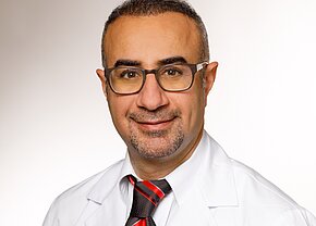 Hamid Al-Sudani Ein freundlicher männlicher Arzt mit Brille und kurzem, grauem Bart lächelt in die Kamera. Er trägt einen weißen Laborkittel und ein gestreiftes, schwarzes und rotes Hemd mit Krawatte. Der Hintergrund ist hell und neutral.