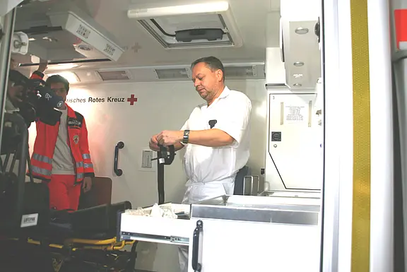 Innenraum eines Rettungswagens: Ein medizinischer Mitarbeiter in Weiß bereitet ein Gerät an der Arbeitsfläche vor. Daneben steht eine Person im roten Einsatzanzug des Deutschen Roten Kreuzes. Behandlungsbereich mit Trage und Schubladen sichtbar.