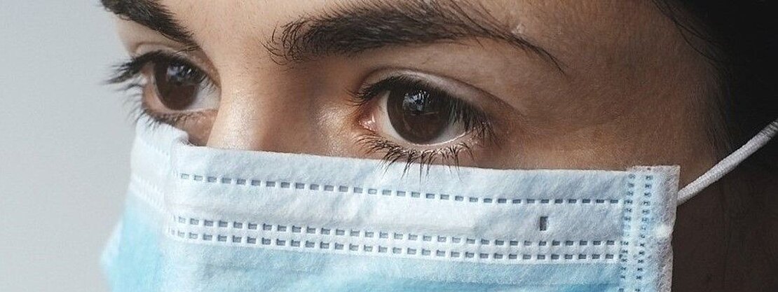 Nahaufnahme einer Person mit blauem medizinischen Mund-Nasen-Schutz; der Blick geht zur Seite, die Augen sind deutlich sichtbar und betonen das Thema Gesundheitsschutz.