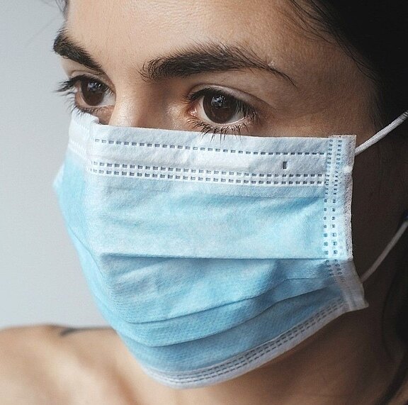 Nahaufnahme einer Person mit blauem medizinischen Mund-Nasen-Schutz; der Blick geht zur Seite, die Augen sind deutlich sichtbar und betonen das Thema Gesundheitsschutz.