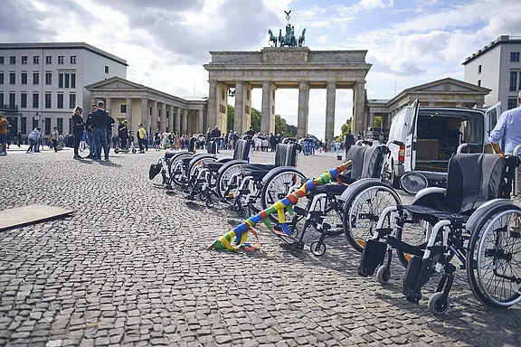 Reihe leerer Rollstühle auf einem kopfsteingepflasterten Platz vor dem Brandenburger Tor. Menschen im Hintergrund; ein bunter Hindernisstab und ein Transporter deuten auf eine Aktion oder Veranstaltung zur Barrierefreiheit hin.