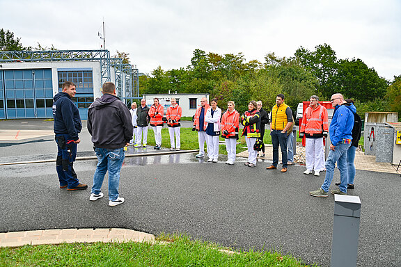 Eine Gruppe von Menschen in Arbeitskleidung steht im Freien. Einige tragen orangefarbene Jacken, andere weiße Kleidung. Sie hören einem Mann zu, der mit ihnen spricht. Im Hintergrund sind Bäume und ein Gebäude sichtbar, das der Gruppe als Kulisse dient.