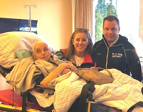 Ein älterer Mann liegt in einem Bett, umgeben von Decken. Neben ihm stehen eine Frau und ein Mann, die lächeln. Die Umgebung wirkt freundlich und hell, mit einem Fenster im Hintergrund.