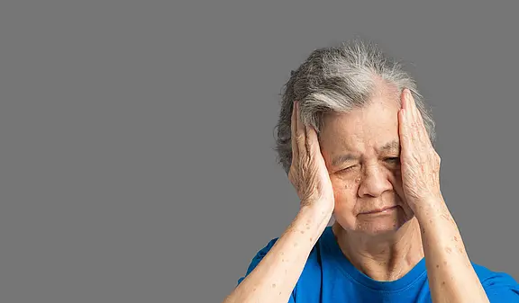 Eine ältere Frau mit grauen Haaren trägt ein blaues T-Shirt. Sie hält sich die Hände an den Kopf und hat die Augen geschlossen, was auf Schmerzen oder Stress hindeutet. Der Hintergrund ist grau und neutral gehalten.