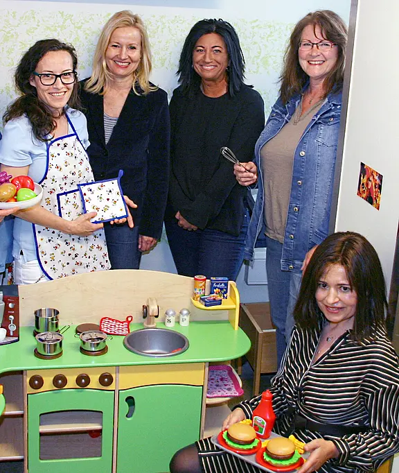 Fünf Frauen stehen und hocken um eine bunte Kinderküche, lächeln in die Kamera. Eine trägt eine Schürze; eine andere hält eine Zange; vorne präsentiert jemand zwei Spiel-Burger mit Ketchup. Auf der Arbeitsfläche liegen Töpfe, Pfannen und Spiellebensmittel.