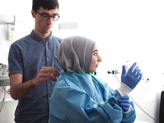 In einem Klinikraum zieht eine Person einen blauen Schutzkittel und Handschuhe an; eine zweite Person hilft, den Kittel am Rücken zu schließen. Die Szene zeigt die Ankleidung von persönlicher Schutzausrüstung vor einer medizinischen Tätigkeit.