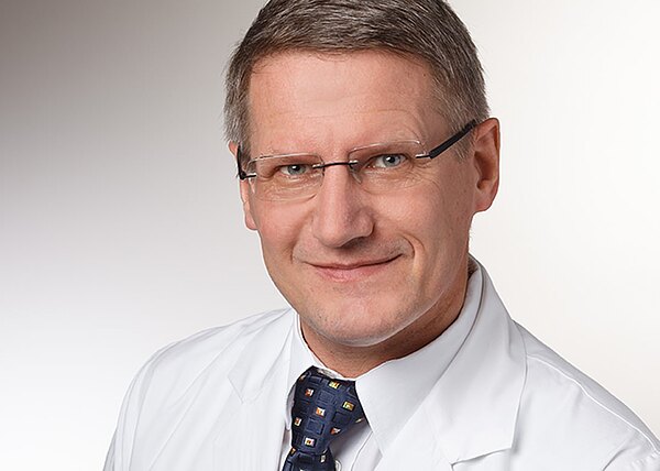 Porträtfoto eines freundlich lächelnden Mannes mit Kurzhaarschnitt und Brille, im weißen Kittel und gemusterter Krawatte, vor neutralem Hintergrund. Er blickt direkt in die Kamera, professionell und vertrauenswürdig.