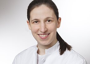 Dr. med. univ. Janina Meladze Kopf-und-Schultern-Porträt einer lächelnden medizinischen Fachkraft mit zurückgebundenen dunklen Haaren, in weißem Kittel, vor neutralem Hintergrund.