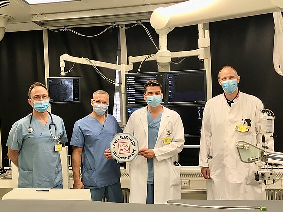 Vier medizinische Fachkräfte stehen in einem Hightech-Behandlungsraum mit Monitoren, tragen OP‑Kleidung und Masken. Der Mann in der Mitte hält ein rundes Zertifikatsschild, das die Abteilung als zertifiziert ausweist.