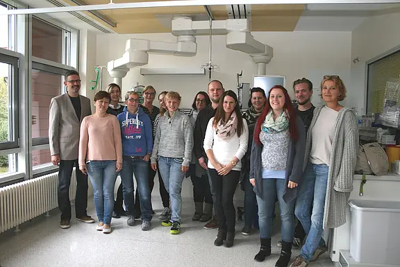 Eine Gruppe von Erwachsenen posiert für ein Gruppenfoto in einem hellen, klinisch wirkenden Raum mit medizinischer Ausstattung. Alle schauen zur Kamera; die Stimmung ist freundlich.