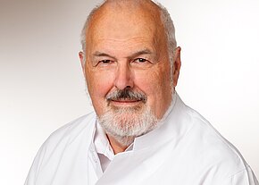 Dr. med. Hans Parsch Porträt eines älteren Mannes mit weißem Haar und Bart, in weißem Kittel, freundlich in die Kamera blickend vor neutralem Hintergrund.