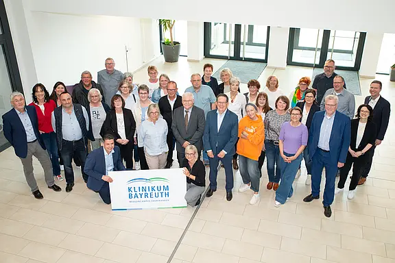 Gruppenfoto von Mitarbeitenden in einer hellen Eingangshalle: Rund 30 Erwachsene stehen lächelnd zusammen; vorn halten zwei ein Schild mit der Aufschrift „Klinikum Bayreuth“. Die Szene vermittelt Teamzusammenhalt bzw. ein gemeinsames Treffen.