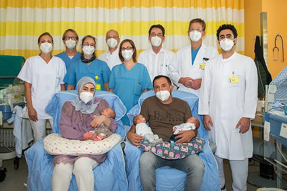 Im Krankenhaus sitzen zwei Eltern auf Sesseln und halten ihre neugeborenen Zwillinge. Hinter ihnen steht ein Team aus Ärztinnen, Ärzten und Pflegekräften in Kitteln. Alle tragen Schutzmasken. Die Szene wirkt wie ein offizielles Gruppenfoto nach einer Geburt.