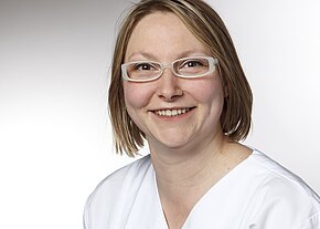Station 25 Porträtaufnahme einer lächelnden Person mit kurz blondem Haar und weißer Brille. Sie trägt weiße Arbeitskleidung, wirkt freundlich und blickt direkt in die Kamera. Neutraler, heller Hintergrund.