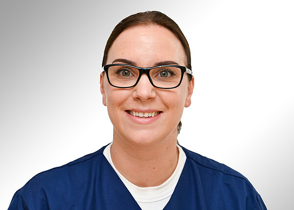 Porträt einer lächelnden Frau in blauer medizinischer Arbeitskleidung. Sie trägt eine schwarze Brille und hat die Haare zurückgebunden; neutraler, heller Hintergrund.