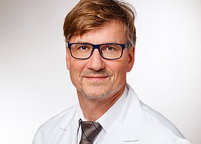 Ein mann in einer weißen Arztkittel und einer braunen Krawatte lächelt freundlich. Er trägt eine Brille mit blauen Rahmen und hat kurze, braune Haare. Der Hintergrund ist hell und neutral, was den Fokus auf sein Gesicht legt.