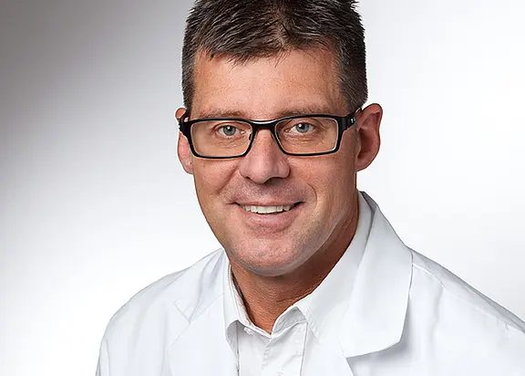 Porträt eines erwachsenen Mannes mit kurzer dunkler Frisur und schwarzer Brille. Er lächelt und trägt einen weißen Kittel vor einem neutralen, hellgrauen Hintergrund.