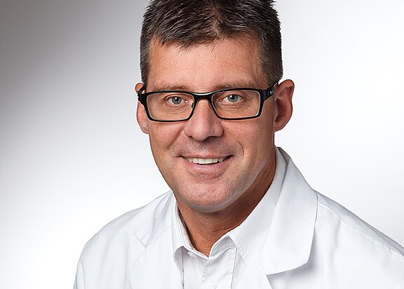 Porträt eines erwachsenen Mannes mit kurzer dunkler Frisur und schwarzer Brille. Er lächelt und trägt einen weißen Kittel vor einem neutralen, hellgrauen Hintergrund.