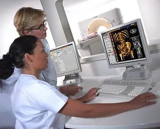 Zwei medizinische Fachkräfte, eine sitzend und eine stehend, betrachten gemeinsam einen Computerbildschirm mit medizinischen Bilddaten. Der Bildschirm zeigt detaillierte Bilder eines Beckenscans. Beide sind in weißen Kitteln gekleidet und konzentrieren sich auf die Analyse der Bilder.