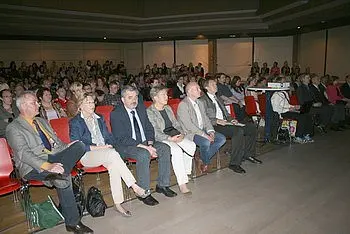 Eine Gruppe von Menschen sitzt auf roten Stühlen in einem Saal. Die Zuschauer schauen aufmerksam in Richtung Bühne, wobei einige Personen im Vordergrund gut zu erkennen sind. Der Raum ist voll mit einer großen Anzahl von Teilnehmern, die an einer Veranstaltung oder Präsentation teilnehmen.