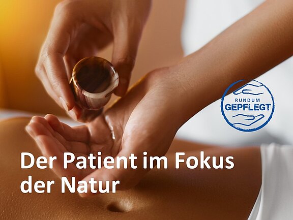 Close-up einer Wellnessbehandlung: Hände gießen Öl aus einem kleinen Gefäß auf den entblößten Rücken einer liegenden Person, Vorbereitung auf eine Massage. Eingeblendeter Slogan: „Der Patient im Fokus der Natur“. Eindruck von natürlicher, sanfter Pflege und Entspannung.