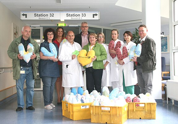 Gruppe von Krankenhausmitarbeitern und Unterstützern im Flur. Einige tragen Kittel. Sie halten bunte, herzförmige Kissen; davor stehen Kisten voller weiterer Kissen. Szene einer Spendenübergabe für Patientinnen und Patienten.