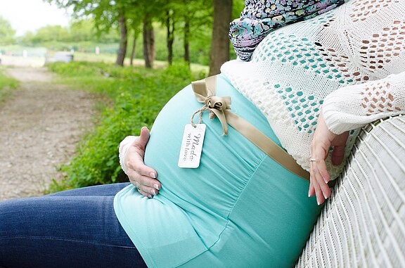 Seitliche Nahaufnahme eines schwangeren Bauchs: Eine Person sitzt draußen, hält liebevoll den Babybauch. Ein beigefarbenes Band mit Schleife und Anhänger mit der Aufschrift „Made with love“ liegt um den Bauch. Stimmung: ruhig, erwartungsvoll.